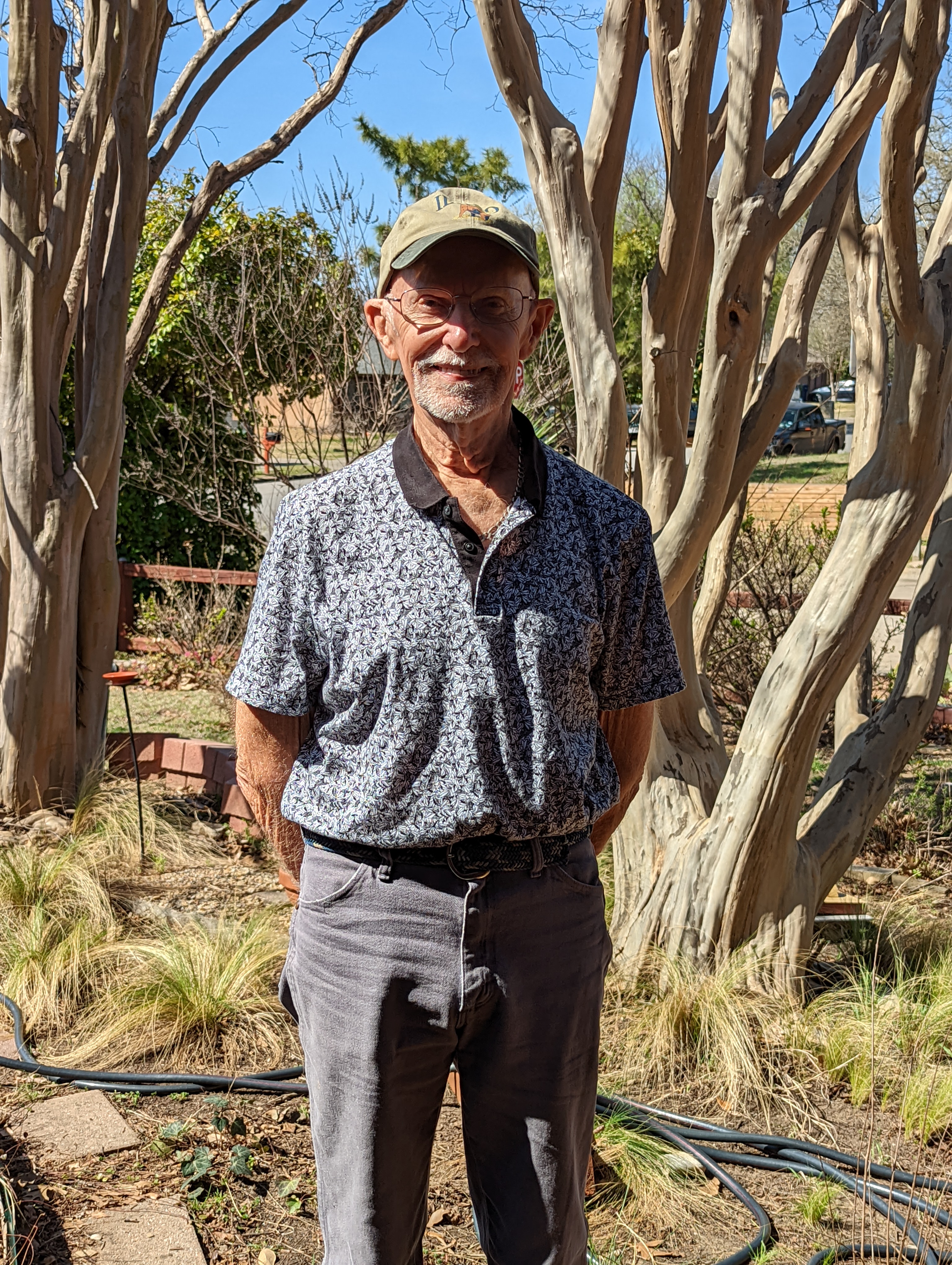 A dapper looking older white gentleman in a ball cap, glasses, a polo shirt, and jeans with his arms behind his back and he is smiling widely in his front yard in Dallas.